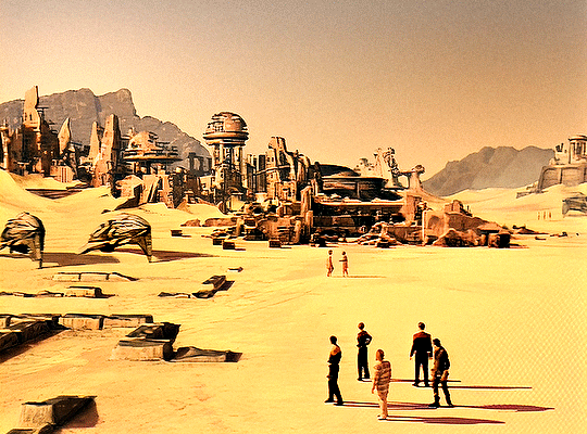 GIF 2: scenic aerial view of a desert planet's surface. a group of 5 people is standin at the bottom of the frame, looking toward the ruins of a city. two people are also running towards the ruins. behind the city is a mountain range.