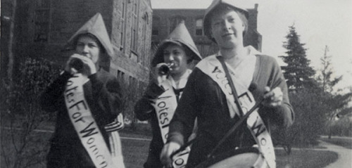 “Students—perhaps members of the Suffrage League—wear ‘Votes for Women’ sashes and march