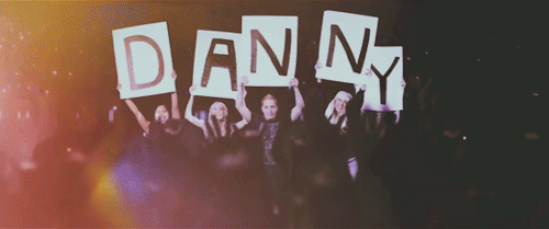 Within an excited audience, five people wave signs which each have a single large letter. Together they spell "Danny".