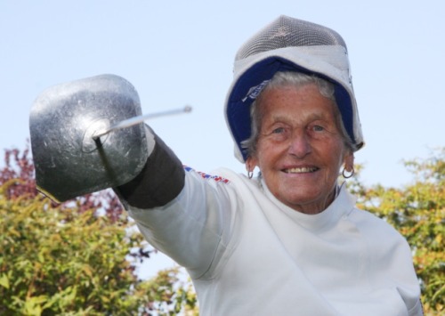 modernfencing: [ID: a veteran fencer in whites with her mask flipped up, aiming her sabre at the cam