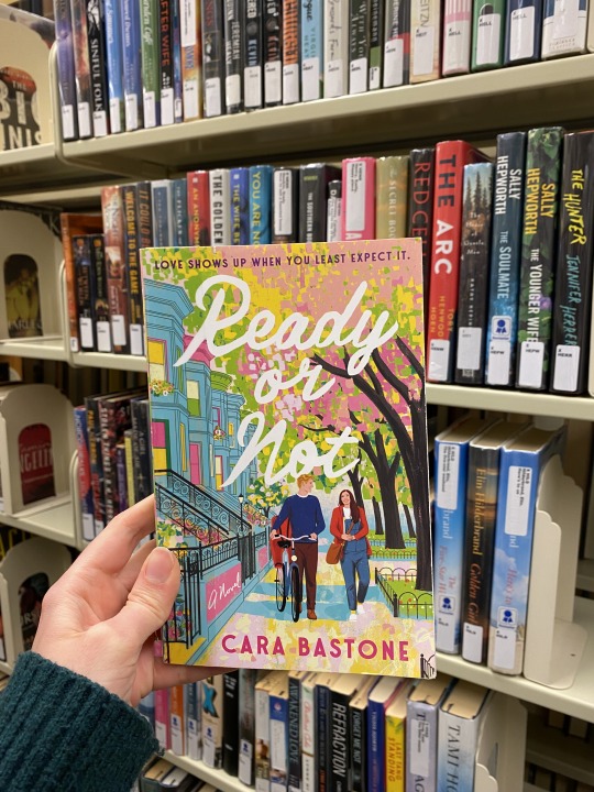 The book held up in front of a shelf of books. Cover shows a brightly colored illustration of a man with a bike walking next to a woman under blossoming trees