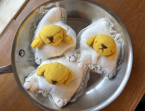 A photo of three plush animals in a metal frying pan. They are made to look like fried eggs; the yolk has a puppy face and ears, while the body is white and amorphous with blobby paws and a lace border. Their faces are simple, with small wide-spaced eyes and a big nose.