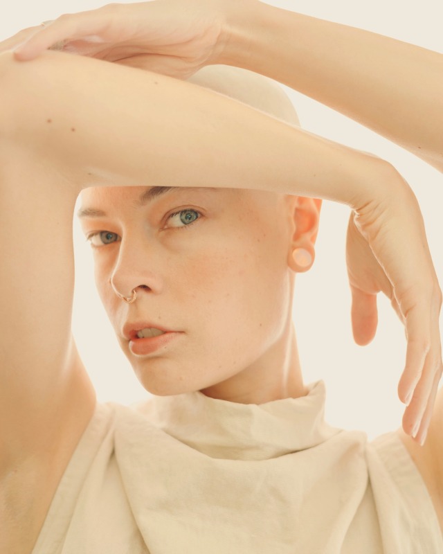 backlit self portrait with arms up around my head. gold septum ring. shaved head. cream linen dress. 