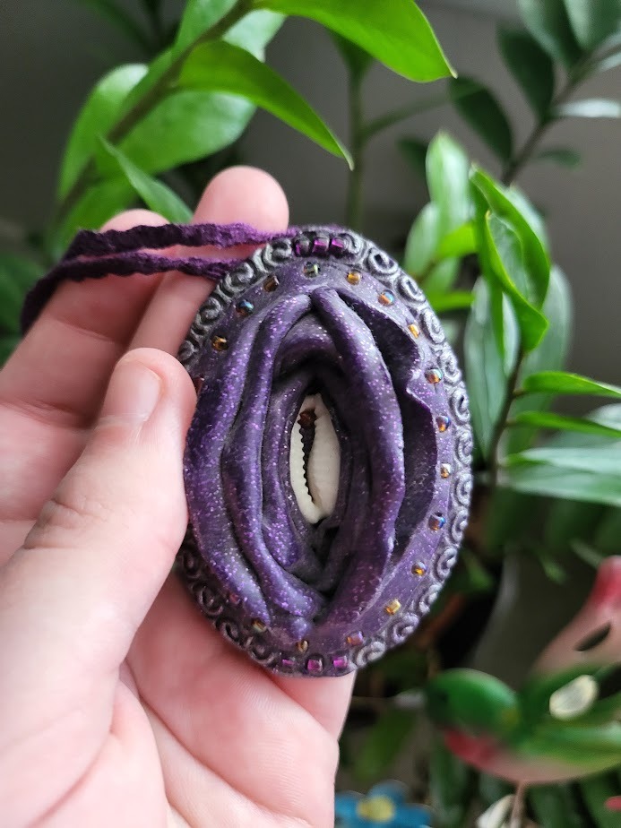 A handmade, purple pendant resembling a vulva with a shell in the center.