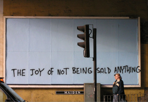 softeningthesound:[Photo ID:Two people walking past a dimly lit white billboard. Graffiti on the bil