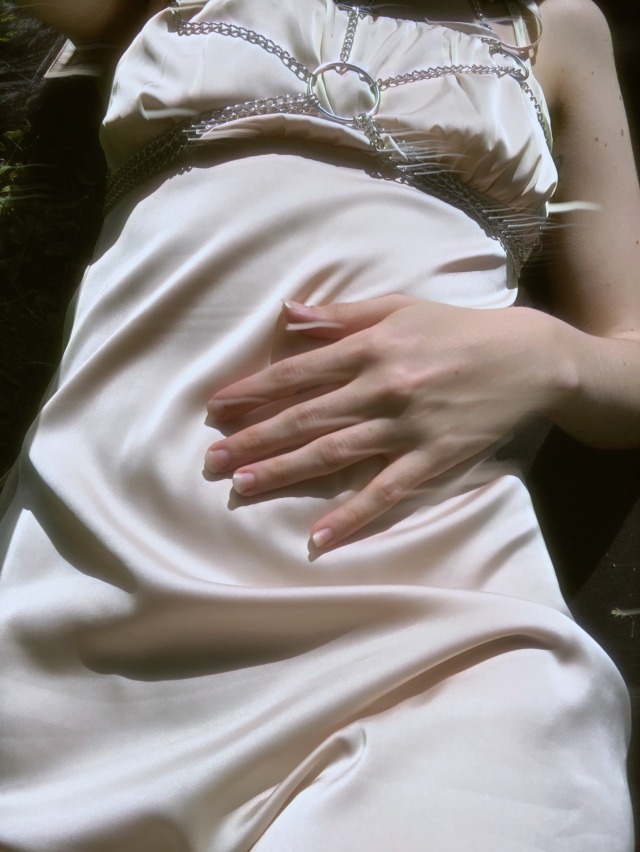 torso selfie, wearing a beige silky dress and silver O ring chain harness, with my right hand cradling my stomach