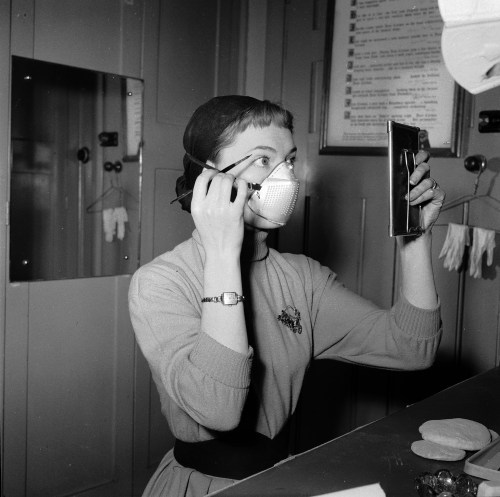 vintageeveryday:A woman putting on makeup while wearing smog mask, London, 1953. Photographed by Jul