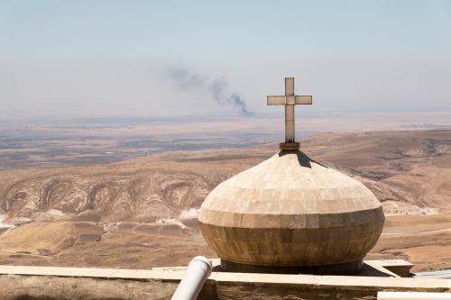 The smoke rising behind this cross is from Mosul, Iraq. The photo was taken last year. There are Chr