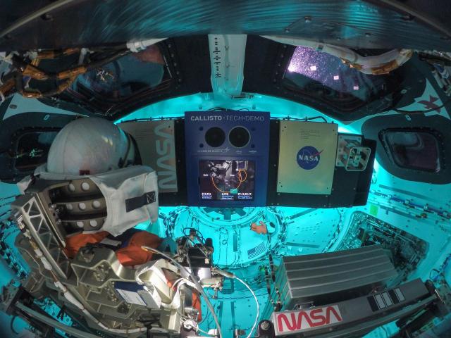 The interior of the Orion spacecraft, bathed in a soft blue light. The back of Commander Moonikin Campos’ head can be seen from behind the commander’s seat. He is wearing an orange Orion Crew Survival System spacesuit and is facing the display of the Callisto payload, Lockheed Martin’s technology demonstration in collaboration with Amazon and Cisco. A Snoopy doll can be seen floating in the background. Credit: NASA