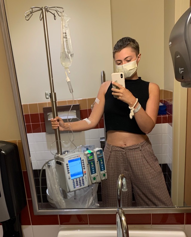 a person with is taking a selfie in a bathroom mirror, they are holding on to an IV pole and have an IV attached to their arm