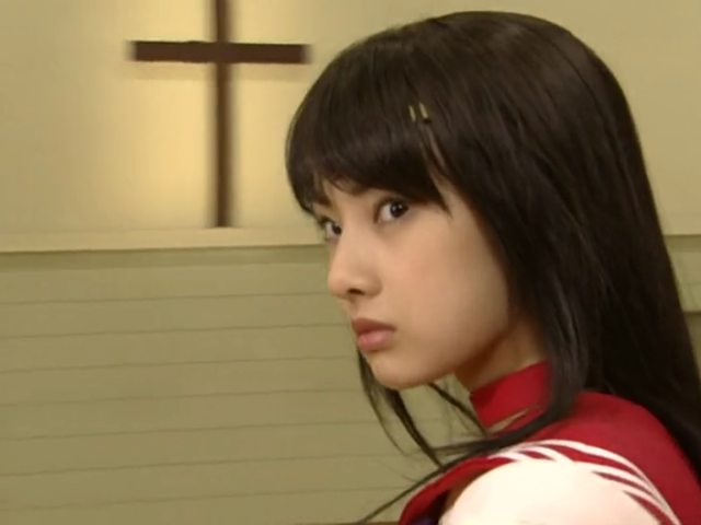 Screencap of Sailor Mars from Toei's 2003 live action Sailor Moon adaptation (played by Keiko Kitagawa) standing with determination. behind her on the wall is a large wooden cross.