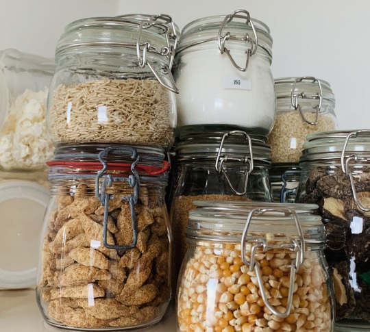 Image shows close up of clean mason jars holding dried staples such as rice, soy puffs, popcorn, dried mushrooms, coconut, MSG.
