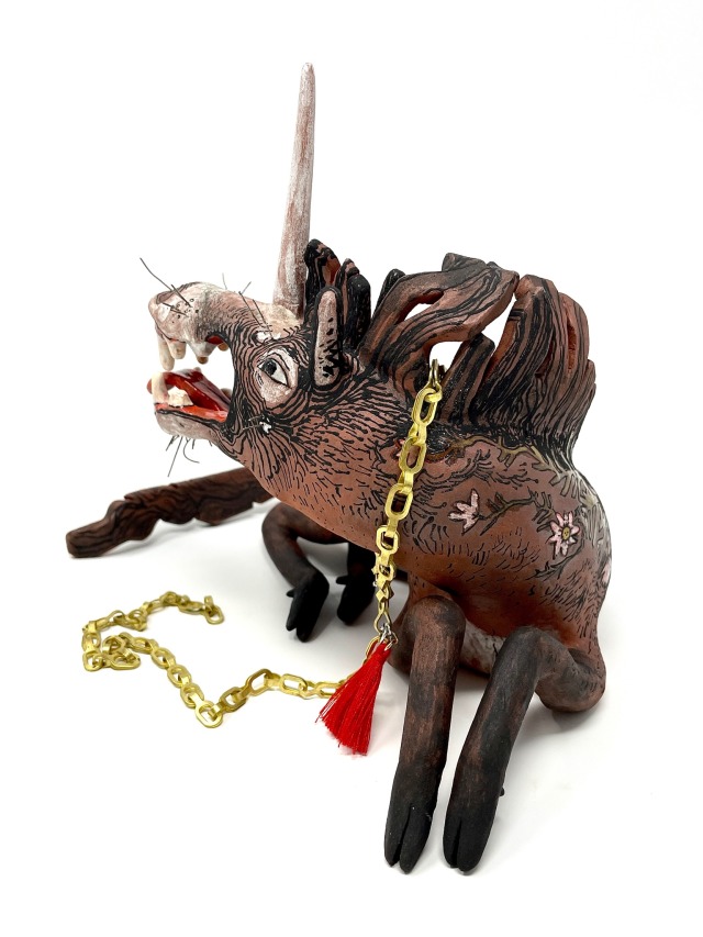 multiple angles of a bay unicorn sculpture resting on all fours, curled into a relaxed half circle, the direction of it’s head and tail completing the arc. delicate cloven hoofs are tucked close to their body, it’s long mane flowing in a breeze.  their maw is open wide, decorated with predacious canine-like teeth, tongue sticking out slightly in a wicked grin that is softened by kind, alert eyes, and wire whiskers frame it’s face. a gold chain with a red tassel is tied around it’s neck and soft pink flowers and leaves decorate it’s backside and sprawl onto it’s neck and rump.