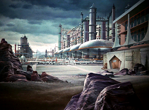 GIF 2: scenic view of an industrial landscape. the foreground is a dirt path with a few rocks scattered around. the background is made up entirely of a large futuristic looking factory stucture. on the right hand side is a building with a pentagon shaped entrance. the sky is filled with dark clouds.