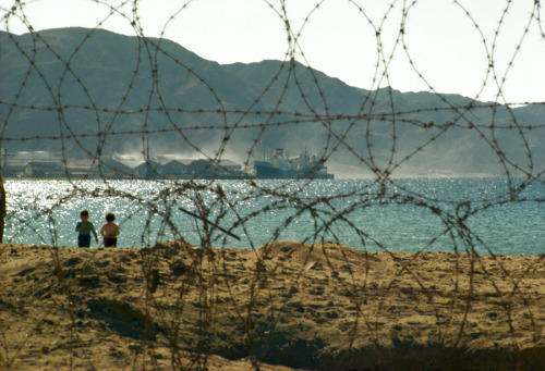 Barbed wire frames view of children on the beach and mountains beyond in Aqaba, Jordan, August 1965.