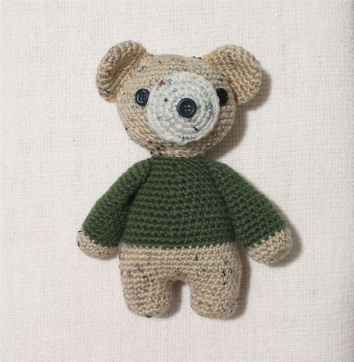 a completed crocheted teddy bear, with button eyes, flecked beige fur, and a green sweater.