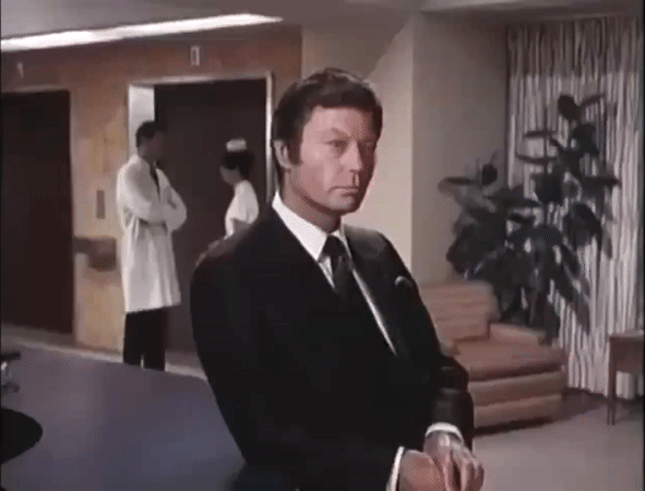 hot older man in a suit stands at the front desk of a hospital and turns his head