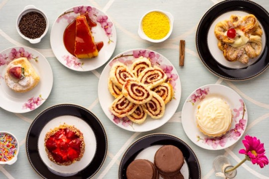 A fancy spread of delectable baked goods, interspersed with bowls of various nonpareils. A small vase with a brightly colored flower is at the lower right.