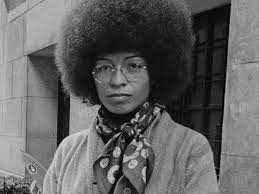 A black-and-white photograph of Davis, a Black woman, with a large Afro and glasses staring at the camera.