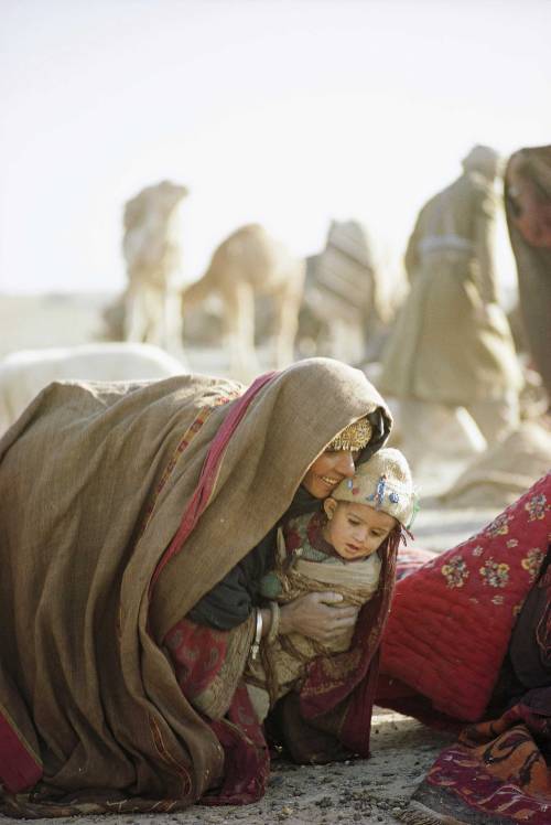 Wrapped against wind and sand, a Kuchi woman lifts a young child. 1968 Source:  Thomas J. Abercrombi