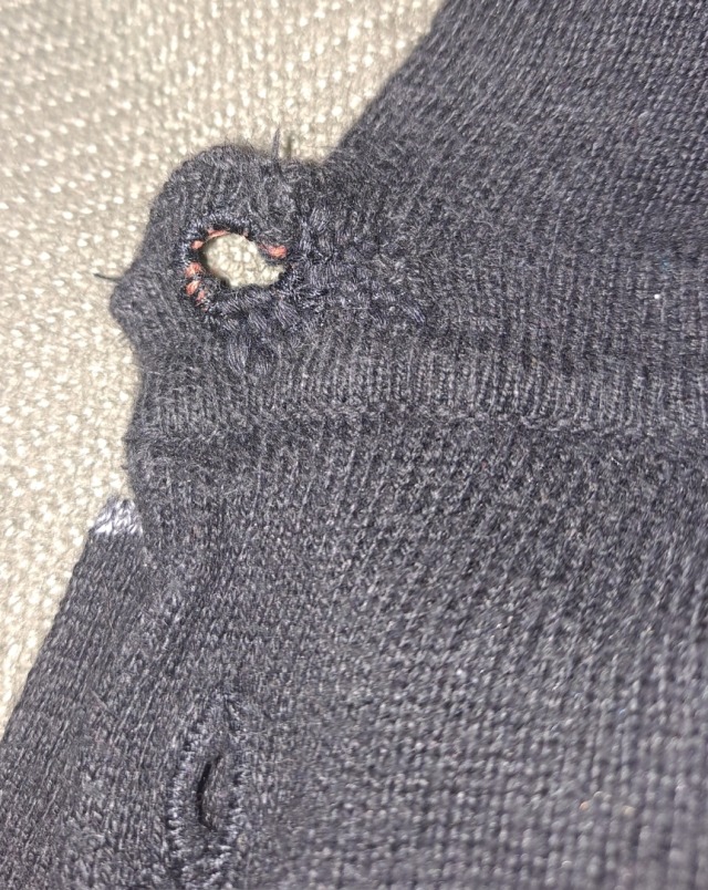 the buttonhole of a black knitted sweater. it is roughly darned with thick black thread and has orange thread peeking out. Green fabric is visible behind the sweater.