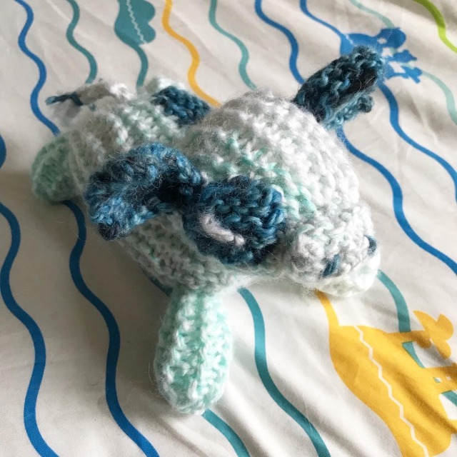A knitted blueberry cow. The cow is a marbled light blue, with dark blue ears and spots. It's eyes are closed, and it sits on a blankee with cartoony ocean waves
