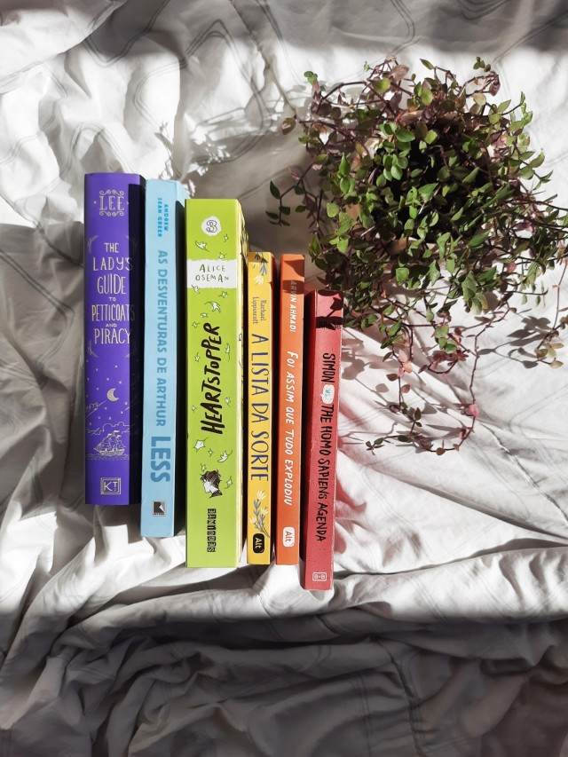 a rainbow book stack showing the spines of the books in a white blanket with a Creeping inchplant in the right side. the books from left to right are the lady's guides to petticoats and piracy, less, heartstopper vol. 3, the lucky list, how it all blew up and Simon vs. The Homo Sapiens Agenda.