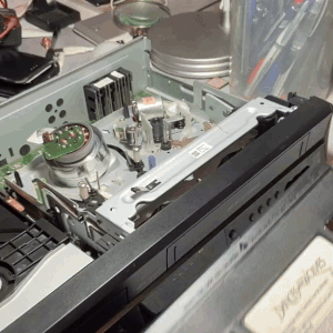 a set of six gifs of a VCR which has had its top removed, so its internal mechanisms can be seen. a person puts a VHS tape in, and we can see the way the tape is pulled out of the cassette and read by the VCR. the person points to the different parts of the machine as it works.