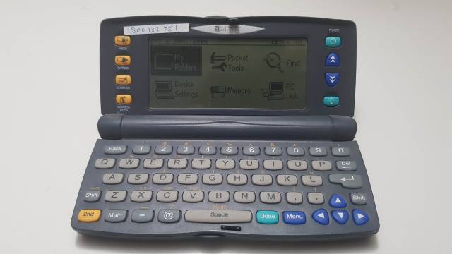 The PocketMail device is open. It resembles a small, handheld laptop computer. The upper portion of the device includes a green and black monochromatic screen with buttons on either side to send and receive emails and navigate pages. The lower portion of the screen contains a simple keyboard for composing emails. The device itself is rounded slightly and has a dark grey colour.