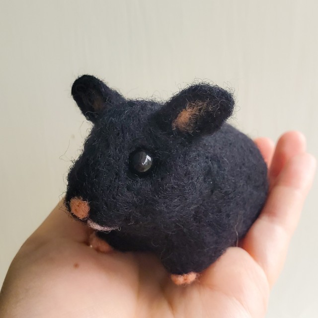 a photo of a needle felted, black hamster. The hamster is being held by a hand against a gray background.