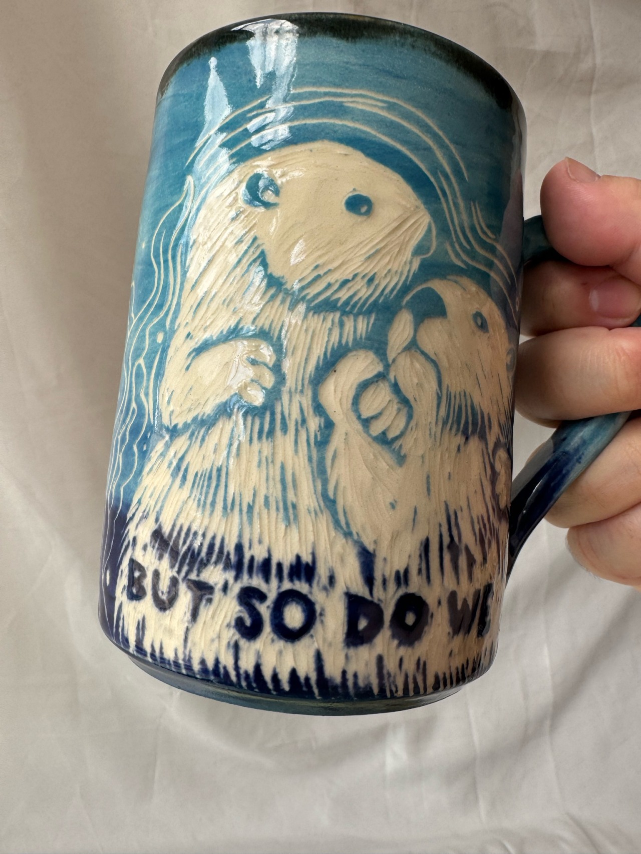 the other side of the mug. it reads 'but so do we' and is decorated with a carving of two otters holding hands