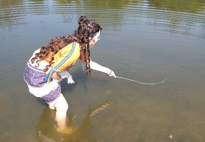 Woman standing in mid-calf-length water. She's seen from the side/back, bent over and looking right. She holds up her skirt with her right hand and stretches her left arm in front of her, holding a thin reed. The tiny bee is barely visible as a small beige lump at the end of the reed.