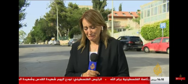 Screenshot from a news broadcast. A news correspondent with long brown hair is looking down and sobbing. She is holding a microphone with the Al-Jazeera logo on it. The background is a neighborhood in Jerusalem. 