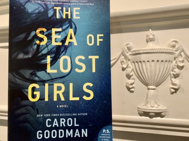 Shown is THE SEA OF LOST GIRLS (with cover art depicting a woman's face under water) beside a carving of an urn. Photo by AHS.