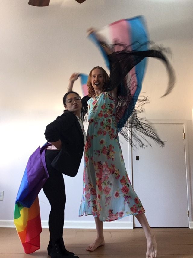 Two Queer cuties bedecked in pride flags have fun together in fun outfits. The tall redheaded nonbinary wears a floral print dress with pearls, a black shawl, and black lipstick as well. Their partner looks sharp in a black sport coat and black velvet leggings, nice shoes too! She's making kissy faces at the camera looking all cute. She's got a Masters Degree in Information Science and I'm so proud of her.