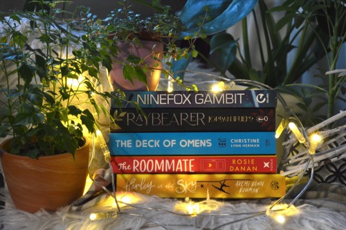 stack of books surrounded by plants, the books are from top to bottom, all in paperback: ninefox gambit, raybearer, the deck of omens, the roommate, harley in the sky