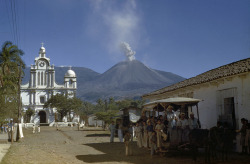 unrar:  People wait in mule-drawn tram; nearby volcano spouts steam and ash, El Salvador, Luis Marden.