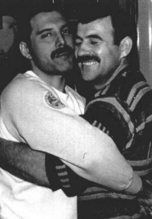 Black-and-white photo of Freddie and Jim, two dark-hair, mustachioed men, embracing and smiling, both wearing soft-looking jumpers. Freddie is looking into the camera, while Jim's eyes are closed.