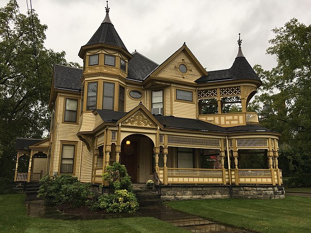 A beige wooden Victorian house with two turrets, long upper and lower balconies, and intricate wooden decorations. It sits on a green lawn surrounded by thin-leafed trees under an overcast sky.