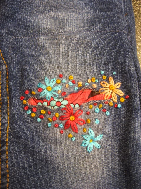 Colourful flower embroidery surrounds a hole in a pair of dark gray jeans. Fabric with a red and black flower print peaks out of the hole.