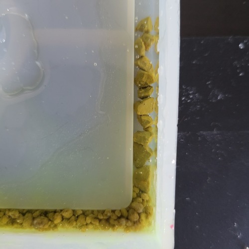 a very close view of a corner of a square silicone coaster mold. Down the channel of the part that will form the coaster ridge, several small, tan rocks are arranged in a tall, thin stack. They are submerged in resin that overflows onto the flat surface of the mold above the channel.