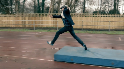 [ID: Five Screencaps Of Dave Gorman On Taskmaster, Traversing An Outdoor Athletics Track With Comically