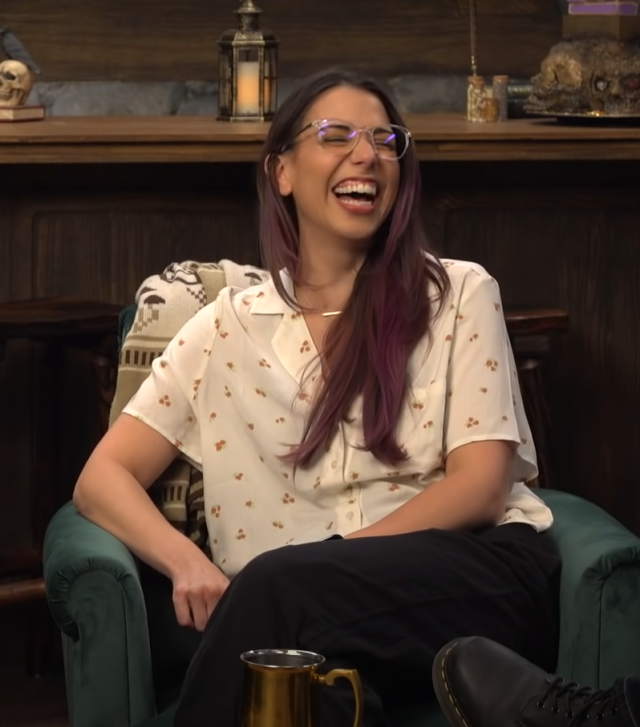 Laura is wearing a cropped white blouse with a small flower pattern on it alone with black pants. Her hair is dark brown with a slightly faded purple red at the ends. She is wearing glasses with clear plastic frames and is laughing at something, her eyes scrunched mostly closed. 