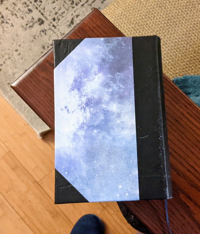 The back cover of a handbound copy of Jules Verne's From the Earth to the Moon and Around the Moon. The spine and corners are covered in black faux leather, and the cover paper has a blue and purple galaxy pattern. 