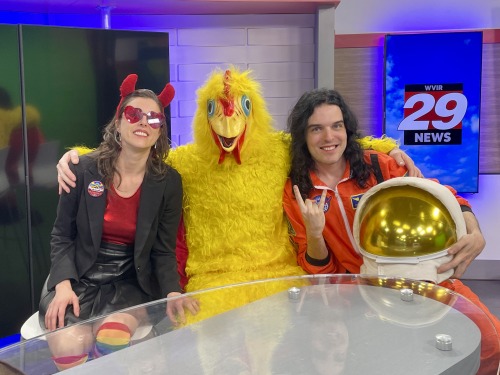 From left to right: Carter Lewis, Lance Brenner, and Hunter Bruton of The Falsies at the NBC29 studio in Charlottesville, VA. Carter is smiling and wearing devil horns, a black skirt and jacket with rainbow socks and a red top, and red heart sunglasses. Lance is wearing his chicken suit and looking at the camera. Hunter is smiling and flashing the sign of the horns while wearing a space suit and holding his helmet.