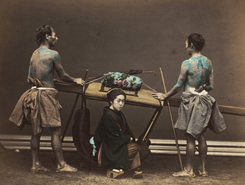 Hand-colored photo of woman resting beside her &lsquo;kago&rsquo; (palanquin carried by men)