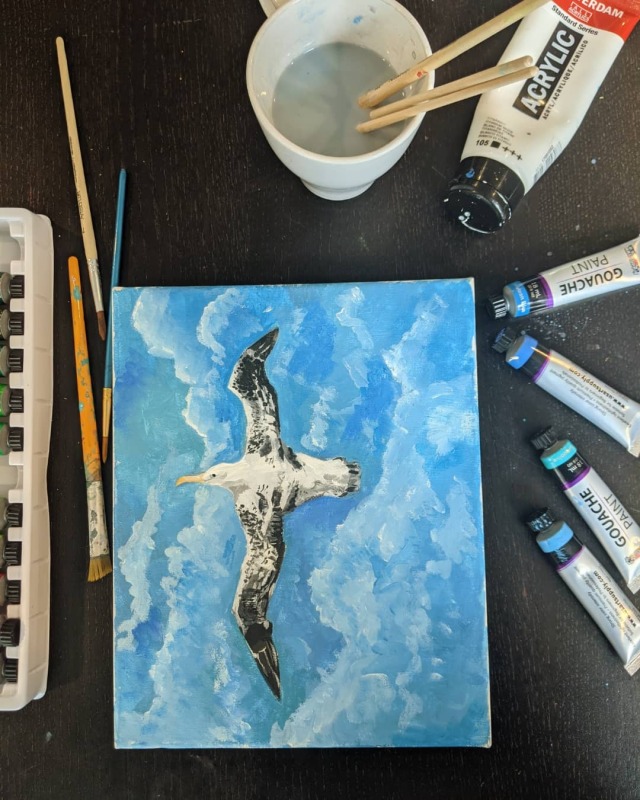 A painting of an albatross in flight over the ocean, surrounded by blue and green paint tubes and mug with paintbrushes and water in it. 