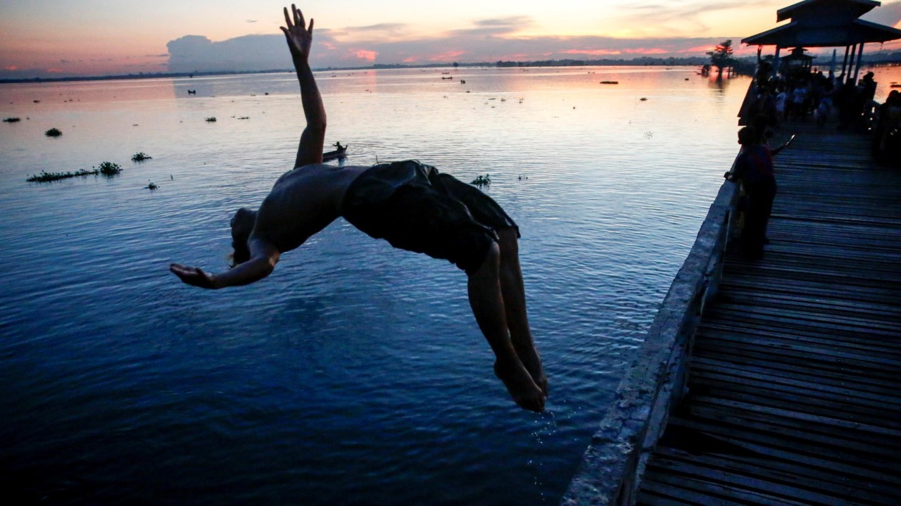 SALTO AL ATARDECER. Los niños de Myanmar saltan al agua mientras la gente disfruta de la puesta de sol en el puente U Pain (U Bein) en Mandalay, Myanmar. La pasarela se extiende 1,2 km sobre el lago Taungthaman conectando sus dos bancos cerca de...