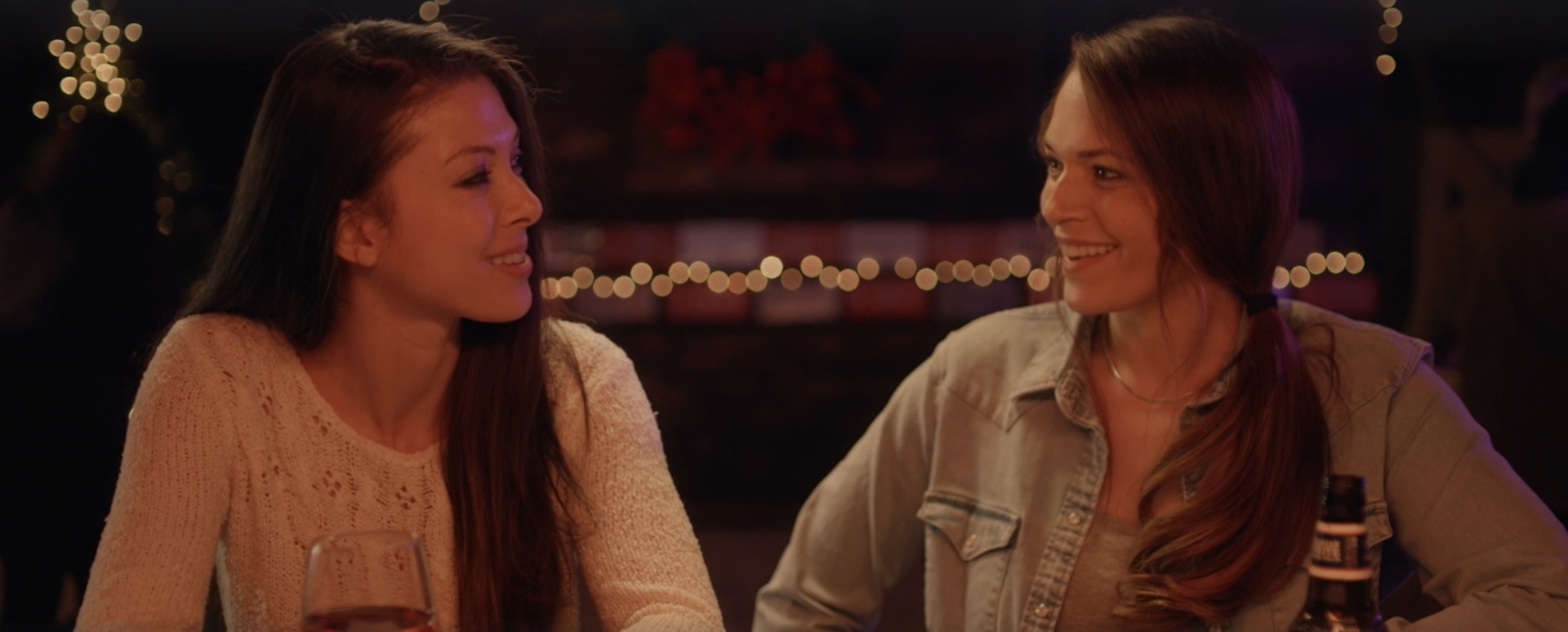 Haleye and Kate from Christmas At The Ranch sit at a bar smiling at each other