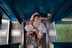 pincuo:  A woman is riding between the railway carriages of a local train heading north from Dhaka, the capital of Bangladesh. Her luggage is tucked under the carriage in front of her. It is the month of Ramadan, a fast which culminates in Eid-ul-Fitr,
